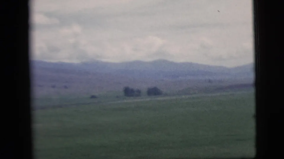 1958 home-movie view of rural fields and distant hills seen through a window, with soft focus and visible 8mm frame edges.