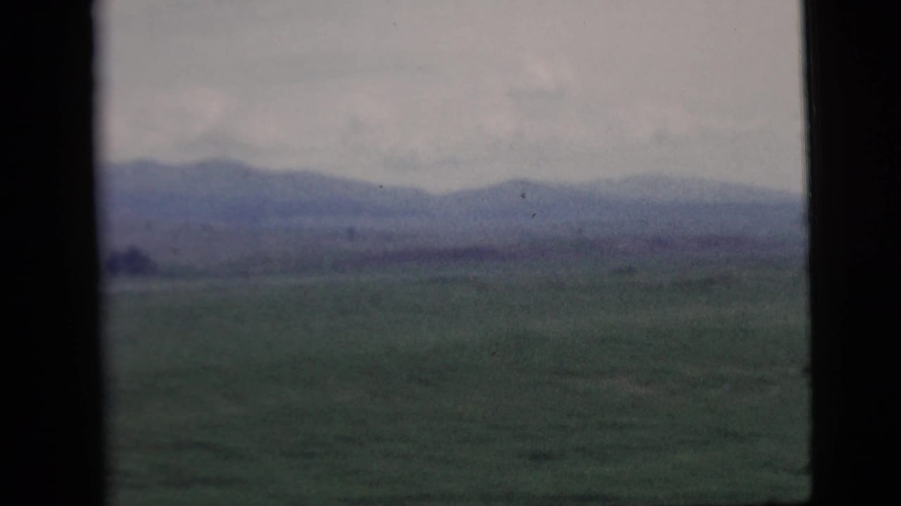 Rural landscape with green fields and distant mountains, viewed through a dark vertical window frame in 1958 8mm footage.