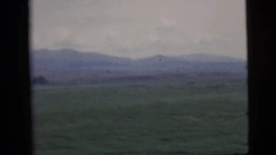 Rural landscape with green fields and distant mountains, viewed through a dark vertical window frame in 1958 8mm footage.