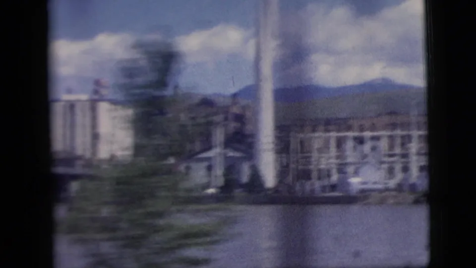 A town or waterfront area with mid-century buildings appears beyond water, blurred by motion and window framing in 1958 8mm footage.