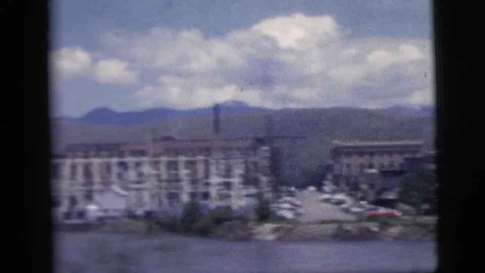 Town or waterfront buildings stand beside water with distant mountains and bright clouds, framed by dark 8mm film edges.