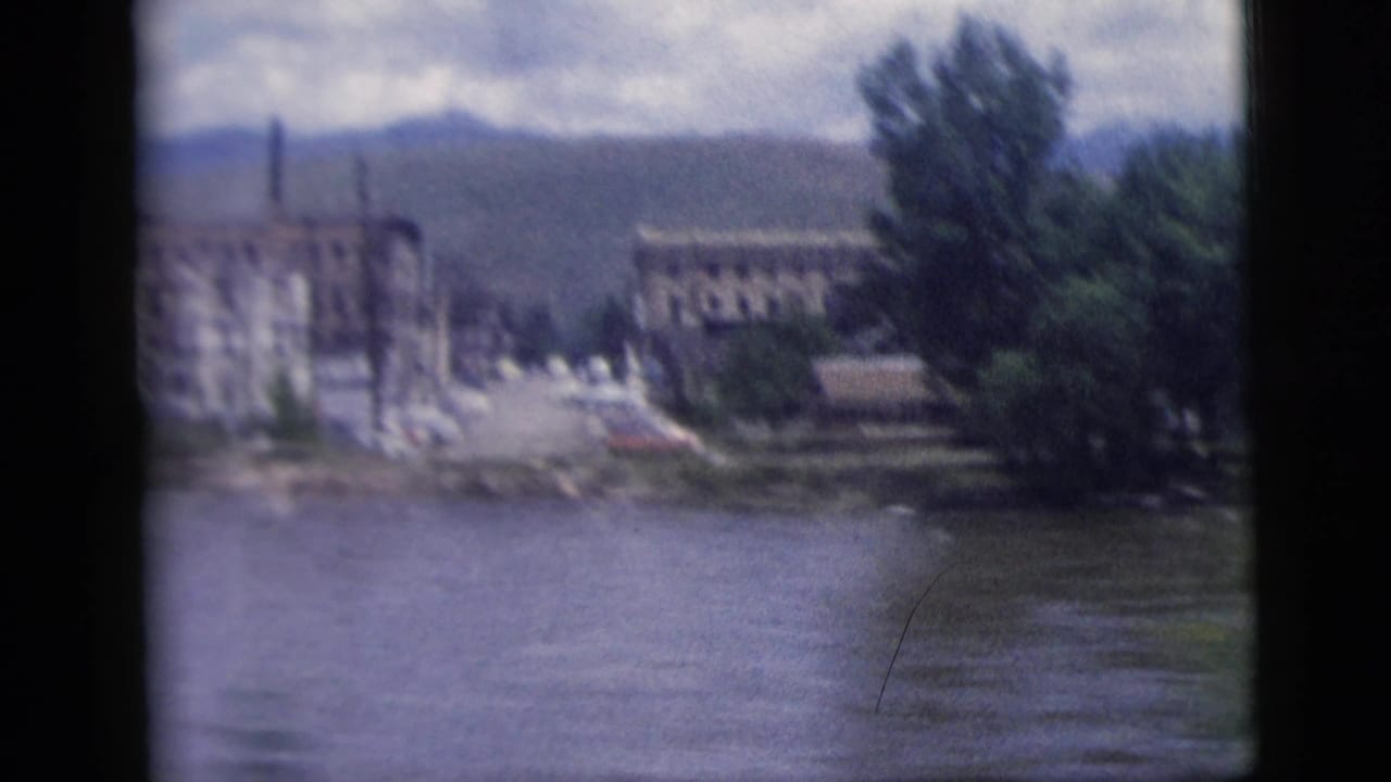 1958 8mm home-movie frame showing a riverbank with large buildings, trees, distant hills, and a cloudy sky from a moving-vehicle viewpoint.