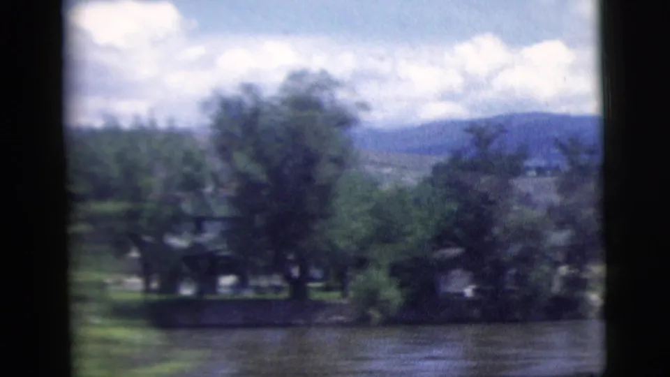 1958 home-movie view across a river toward trees, small shoreline structures, distant hills, and a partly cloudy sky.