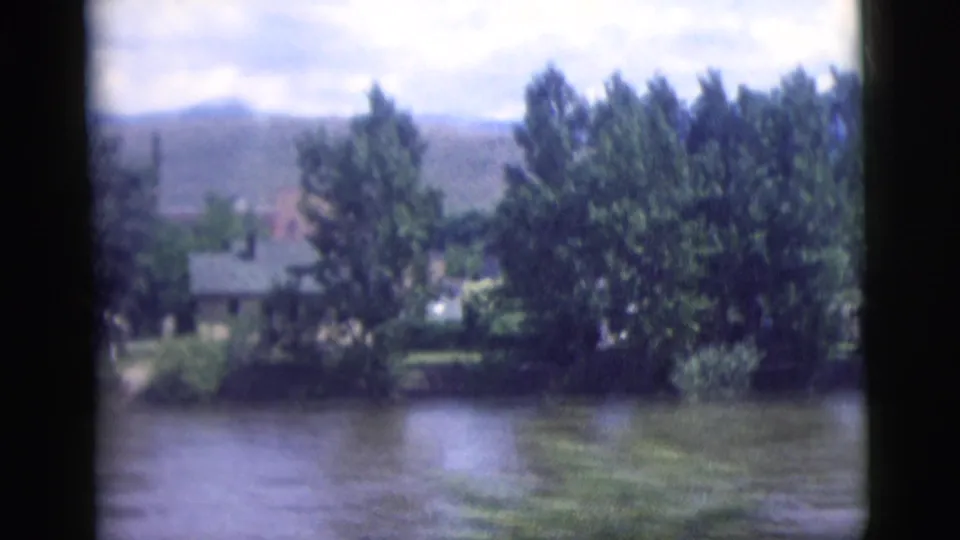 1958 8mm frame showing a riverside house partly hidden by trees, water in the foreground, and hills under a cloudy sky.