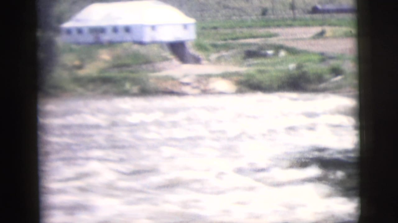 1958 8mm travel frame showing a small white building near a fast river with green terrain behind it.