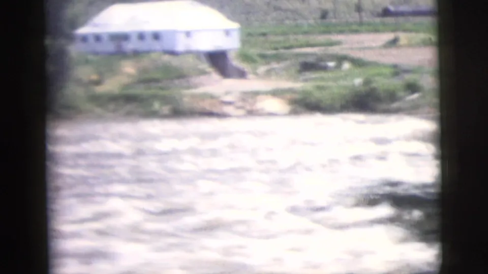 1958 8mm travel frame showing a small white building near a fast river with green terrain behind it.