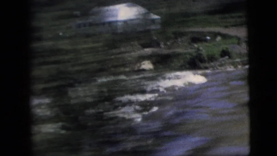 Soft 8mm frame of a river or waterway passing a pale building and dark green shoreline.