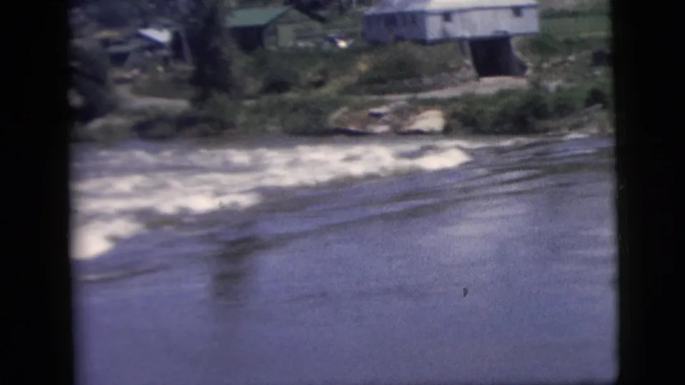 Home-movie travel view of rapids, reflective water, and a small building set back on the riverbank.