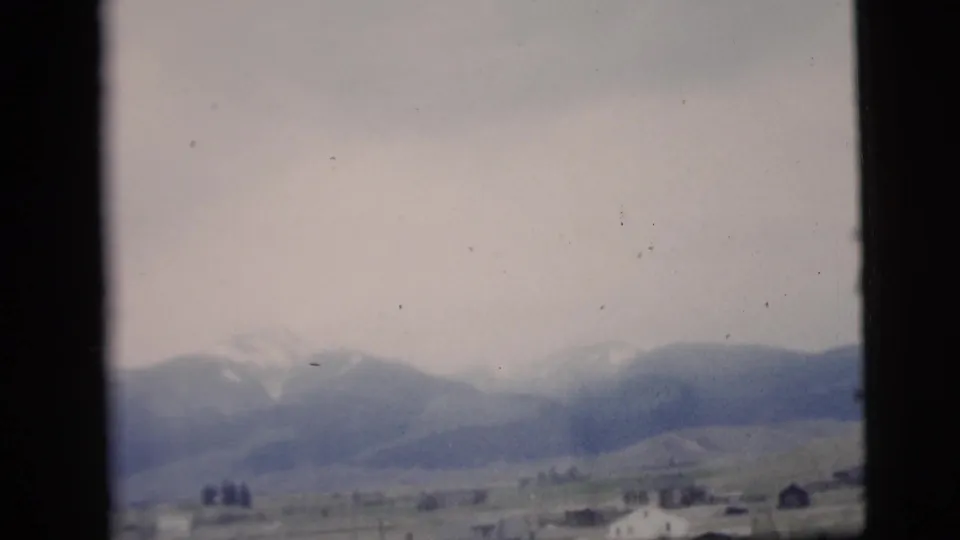 Faded 8mm frame showing distant mountains, pale sky, open ground, and a small foreground structure.