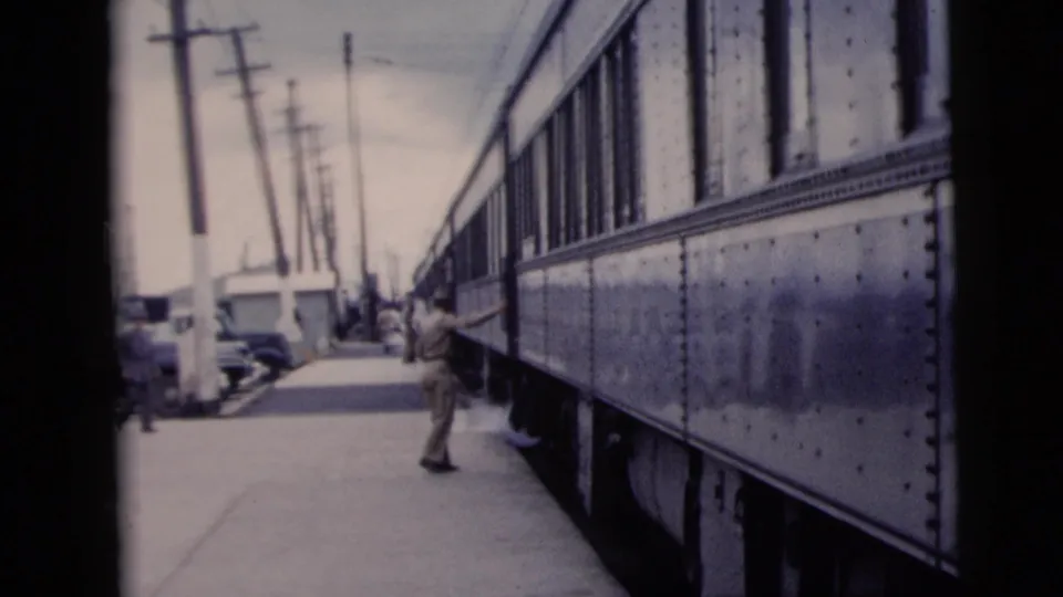 A man in light clothing is seen beside the passenger train on a New York City platform, with poles and parked cars lining the background.