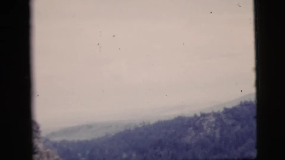 1958 archival home-movie frame showing a hazy mountain ridge under a pale sky, with the lower view partly blocked by a window frame.