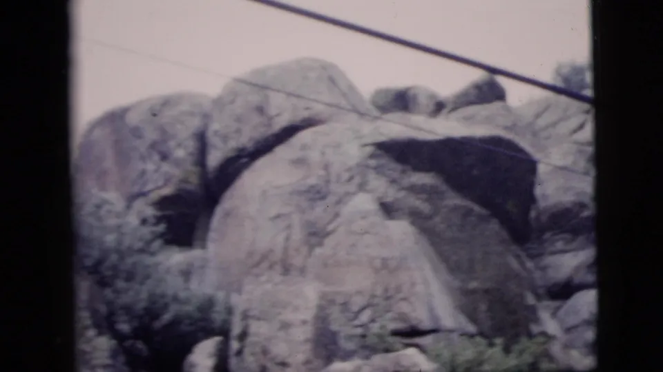 1958 8mm home-movie frame of a rocky hillside with boulders, low shrubs, and a diagonal cable crossing above.