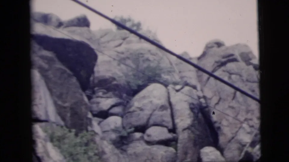 1958 New York City-labeled 8mm view of stacked boulders and shrubs with dark diagonal cables across the scene.