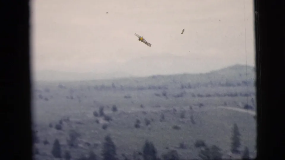 1958 8mm travel frame with a hazy elevated view over a broad landscape or water, with two small aircraft-like specks visible in the sky.