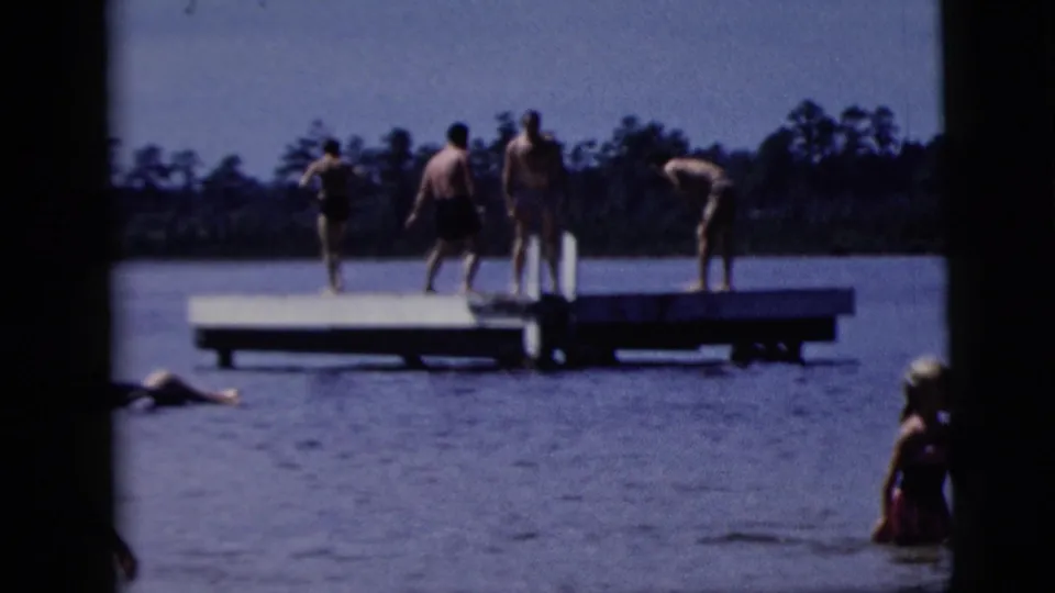 Distant people stand on a low dock in a lake, with swimmers in the water and a dark tree line in the background.