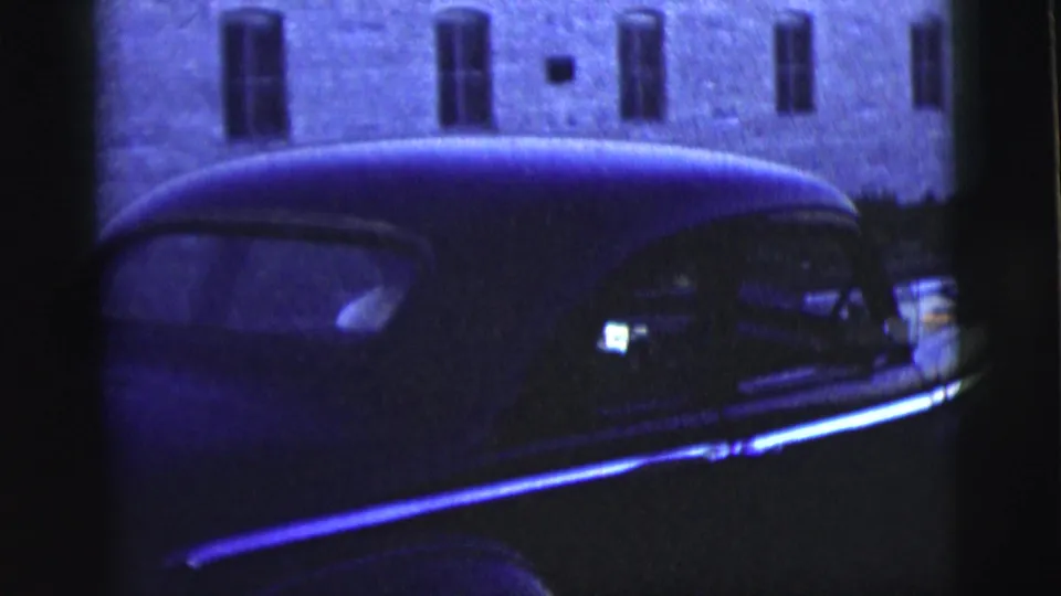 A dark mid-century sedan with chrome side trim appears beside a brick building in 1958 New York City home-movie footage.