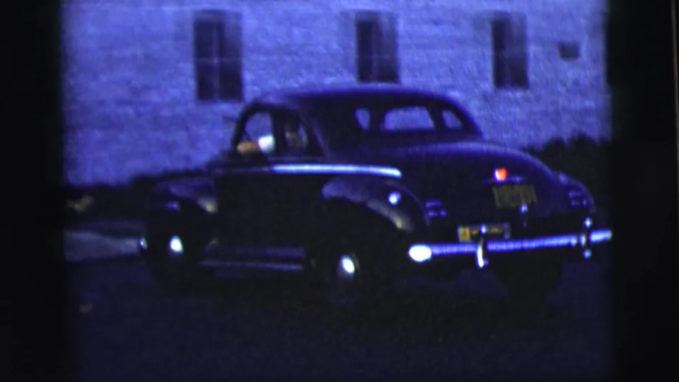 Three-quarter rear view of a dark mid-century sedan with chrome trim and yellow plate in 1958 New York City home-movie footage.