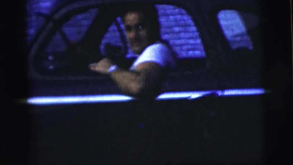 An adult male driver in a white shirt looks toward the camera from inside the sedan during 1958 New York City home-movie footage.