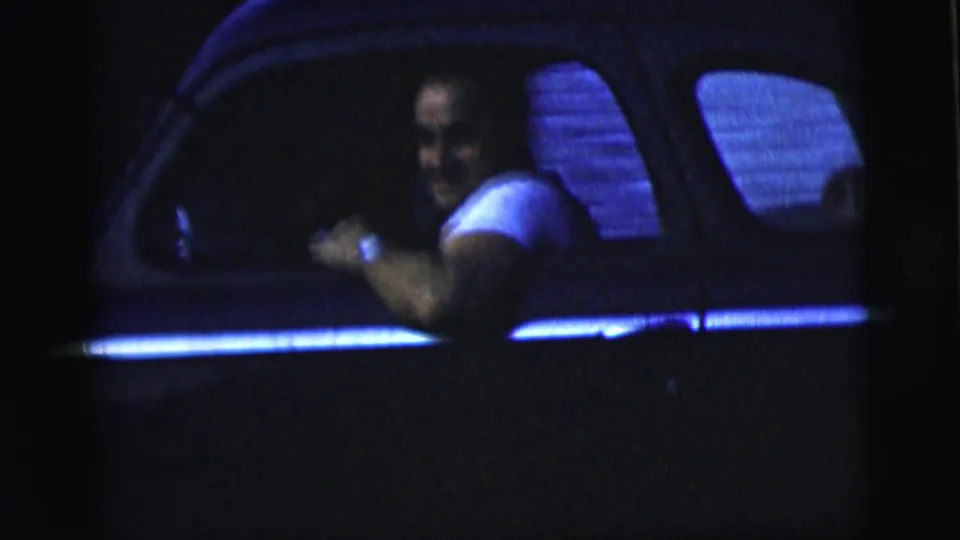 The driver smiles from inside the car, his arm near the open side window in a blue-lit 1958 New York City night scene.