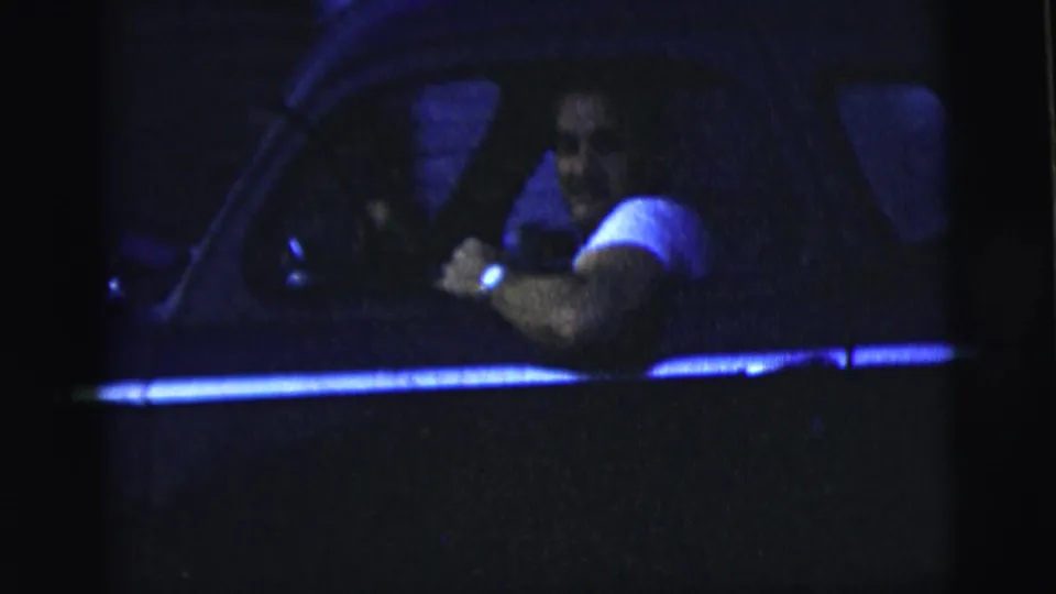 A man driving at night leans near the rolled-down window, with the car interior and door frame visible in soft 8mm grain.