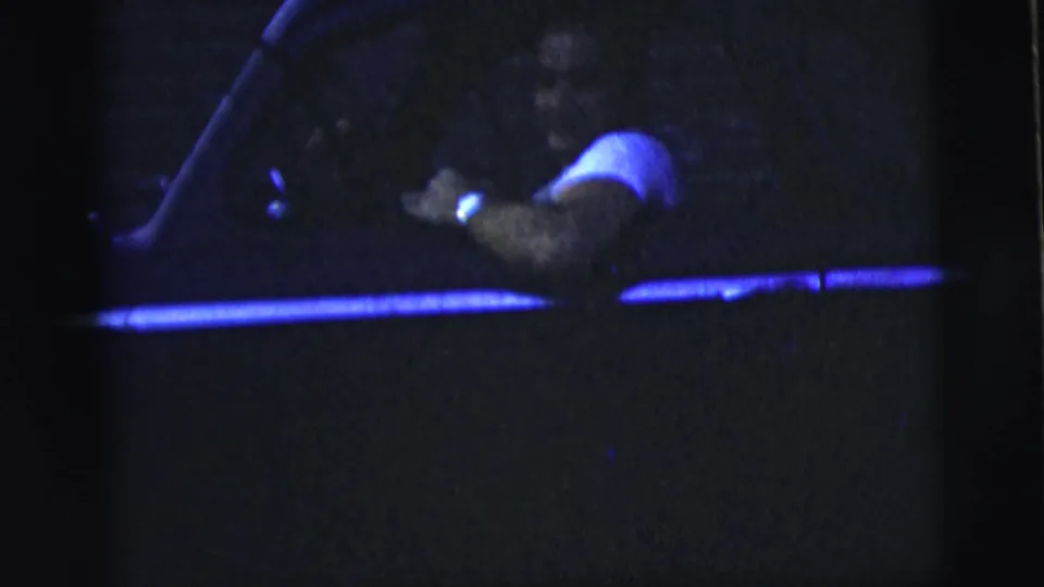 The man remains seated in the dark car, partially obscured by low light as his arm rests near the open window.