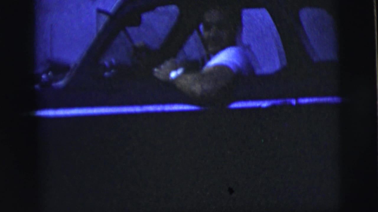 Man in a white shirt driving a dark car at night, seen through the side window.