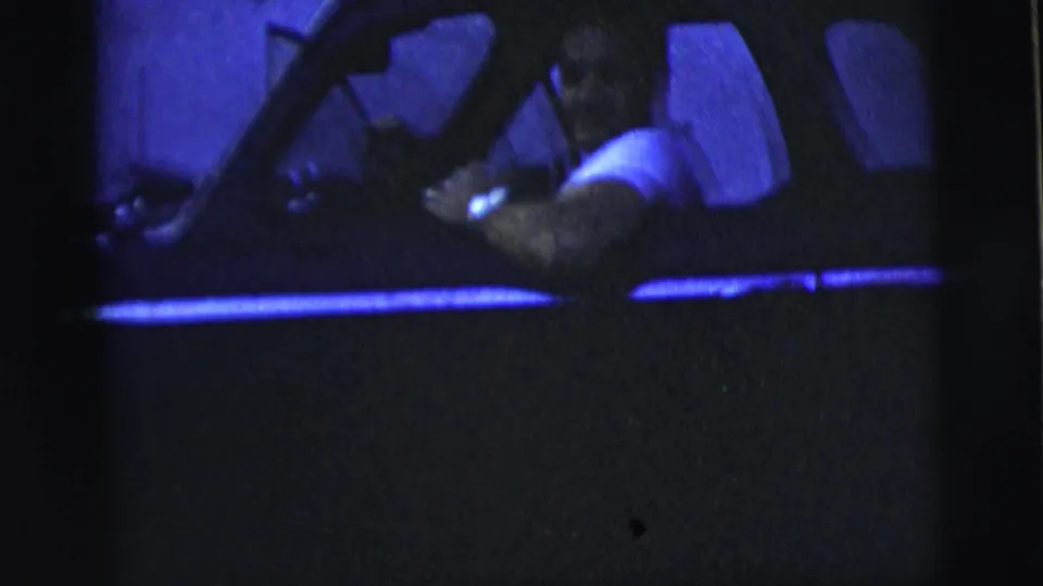 In 1958 New York City home-movie footage, a man in a white shirt drives a dark sedan through low blue night exposure.