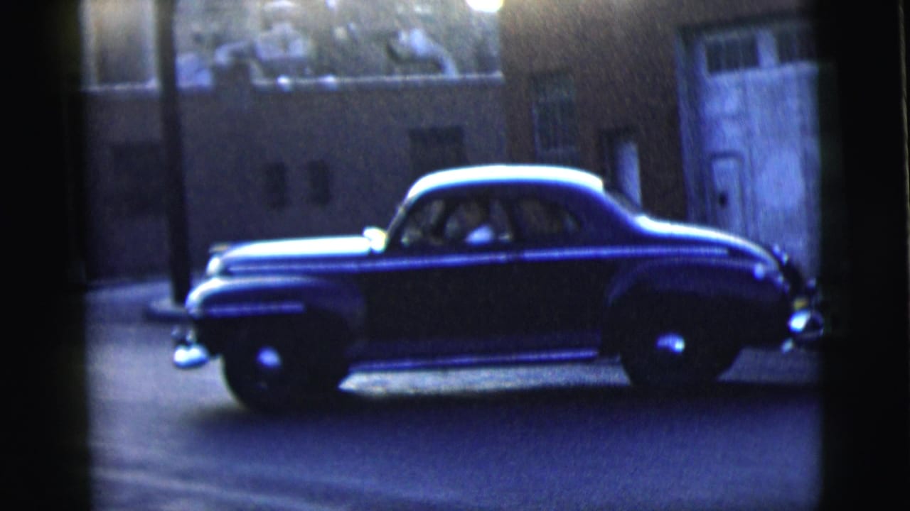 A dark mid-century sedan appears on a dim New York City street at night, with chrome trim catching the available light.