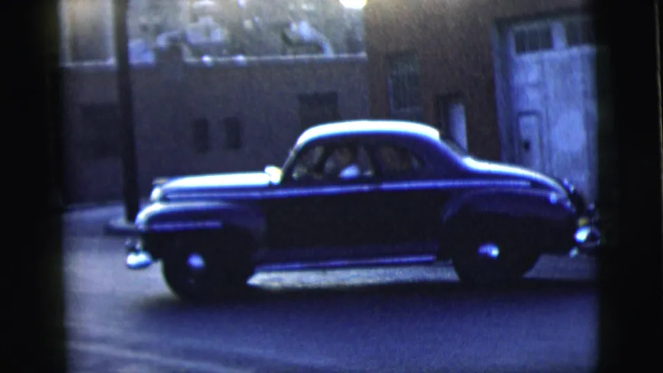 A dark mid-century sedan appears on a dim New York City street at night, with chrome trim catching the available light.