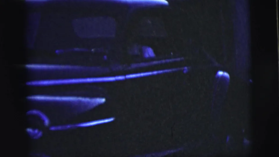 A dark sedan is seen close to the camera in blue night light, with a partial neighboring vehicle shape and indistinct driver area visible.
