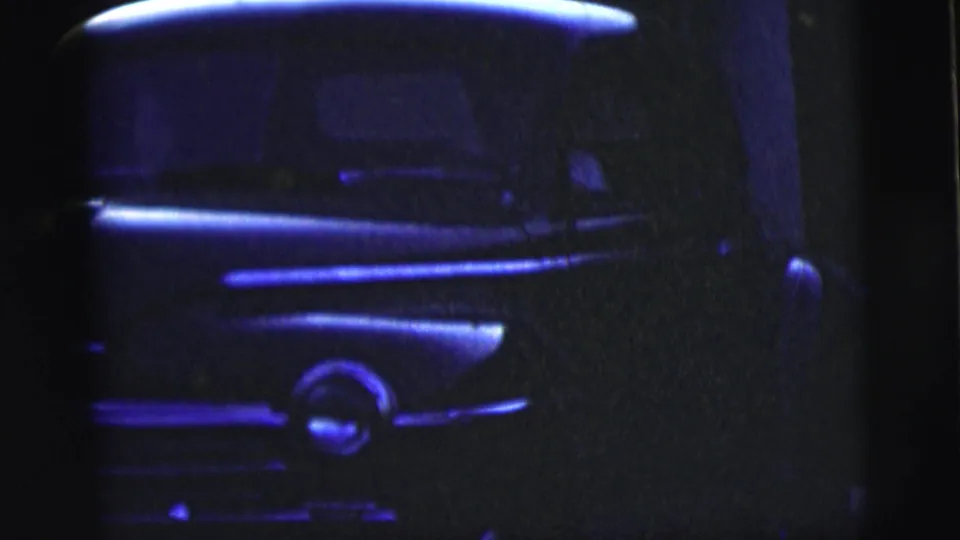 Blue-tinted 1958 8mm frame showing the front end of a classic car, with grille and headlight details partly obscured by shadow.