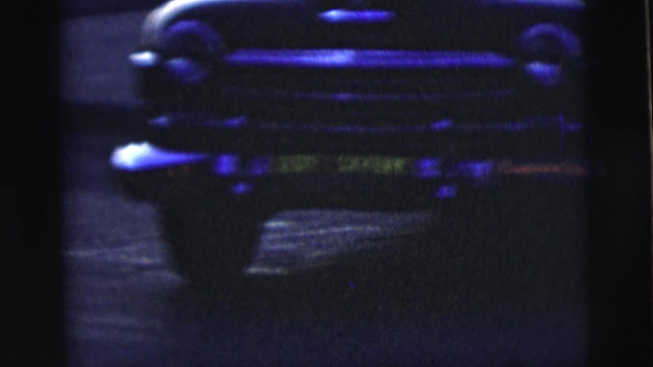 Blue-tinted 1958 8mm frame with a classic car seen from the front, showing rounded hood lines, bumper, grille, and low light detail.