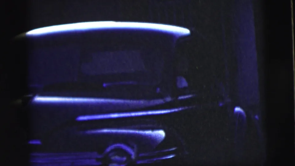 Blue-tinted 1958 8mm frame showing a close view of a classic car's front corner, chrome trim, wheel area, and shadowed bodywork.