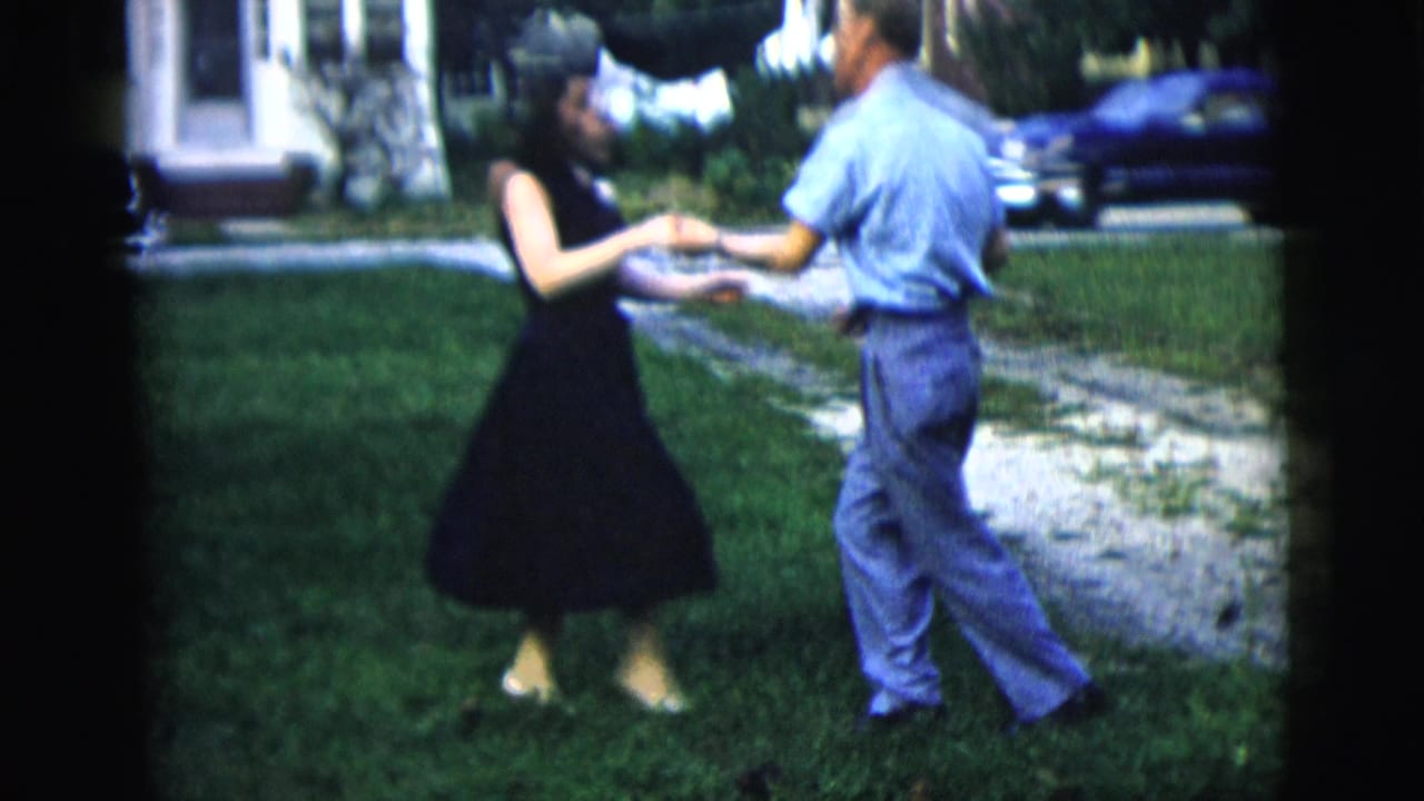 Couple holds hands while dancing on grass near a blue car.