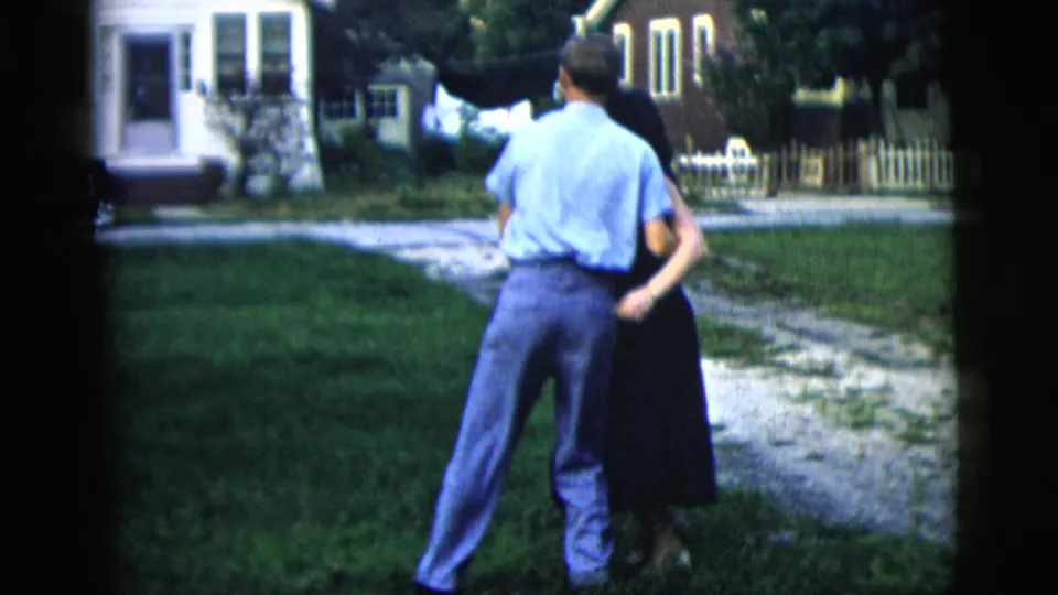 The man is seen from behind as the couple dances closely on the grass, with a driveway, house, and fence in the background.