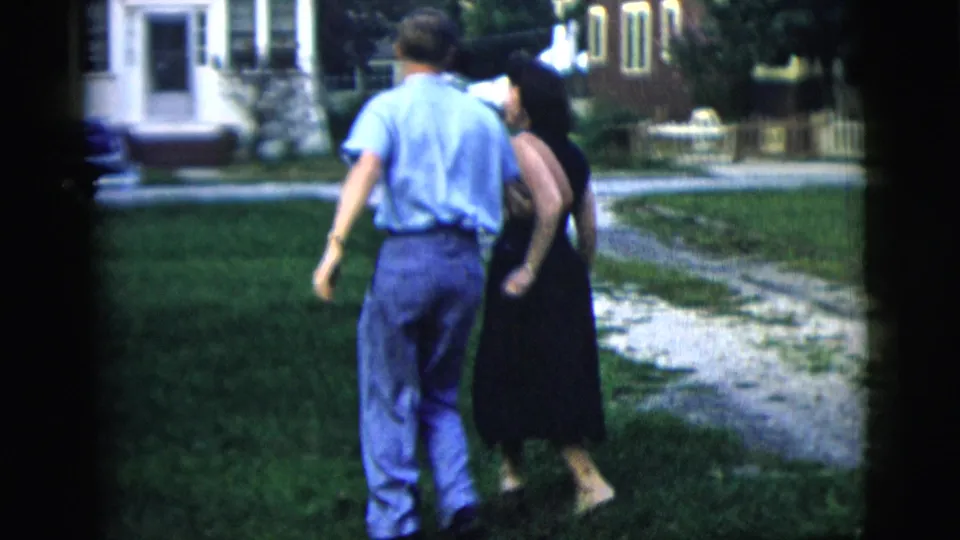 The couple pauses or turns close together on the lawn in 1958 New York home-movie footage, with a walkway and houses behind them.