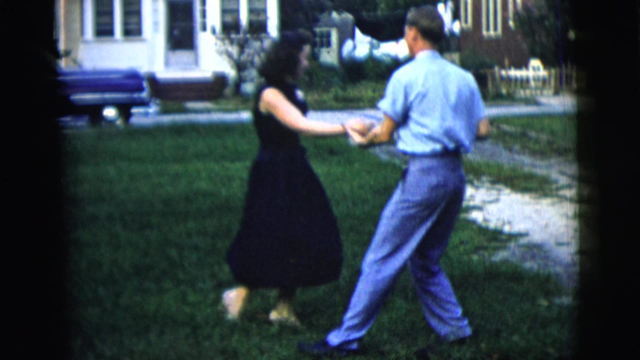 Man and woman hold hands while dancing on a lawn beside a driveway.