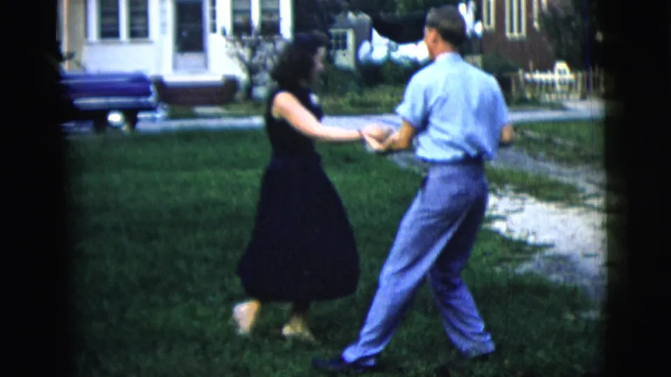 In a 1958 New York home-movie frame, a man and woman dance on a grassy lawn near a house and parked car.