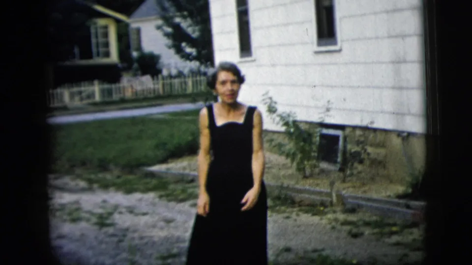 A woman in a black sleeveless dress stands outdoors near a white-sided house, with a yard and fence behind her.