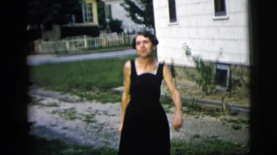 The woman walks toward the camera through a residential yard, with a white house, fence, and neighboring home visible.