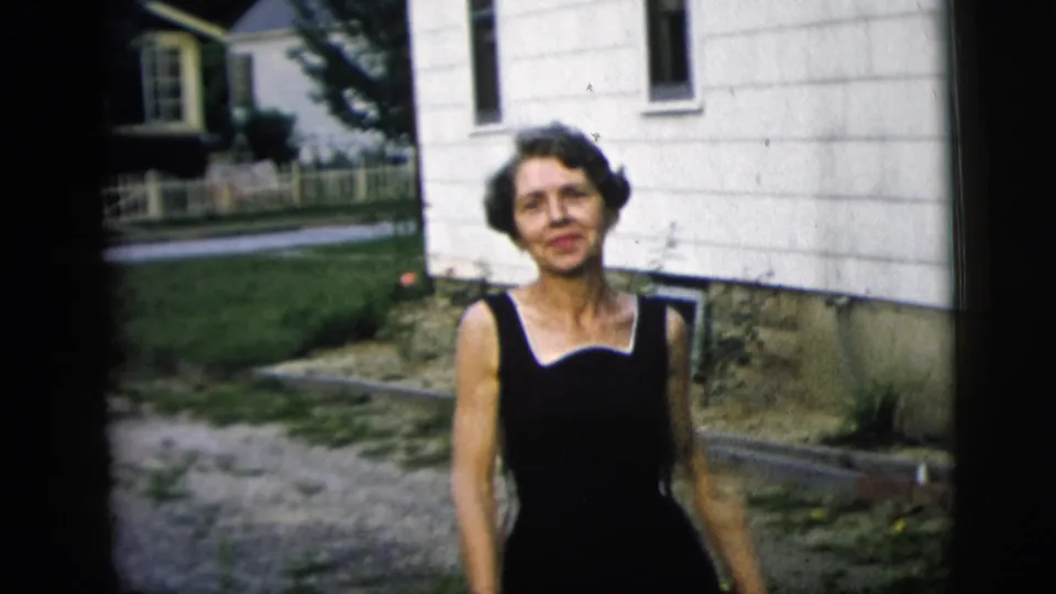 A smiling woman in a black sleeveless dress comes closer to the camera outside a white-sided residential house.