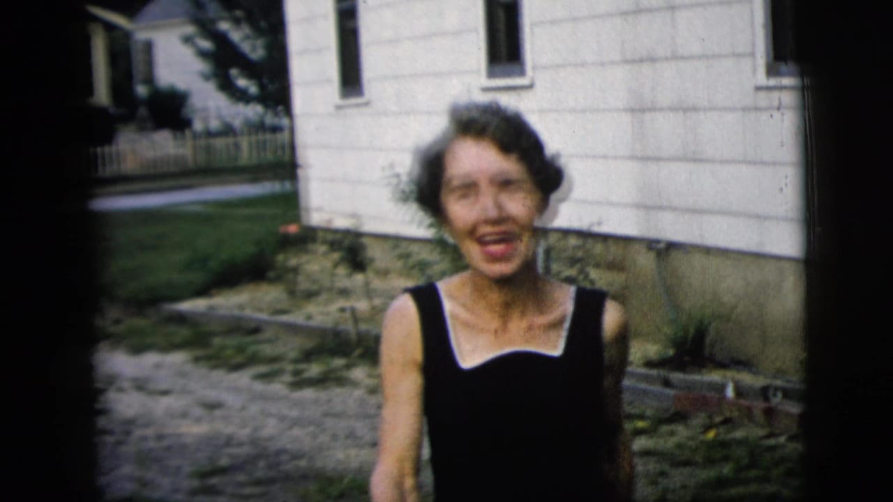 Woman in a black dress smiles near a white-sided house and gravel path.