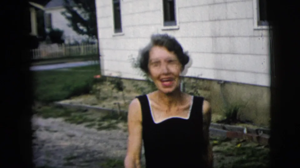 In 1958 New York City home-movie footage, a woman in a black dress smiles toward the camera beside a white-sided house.