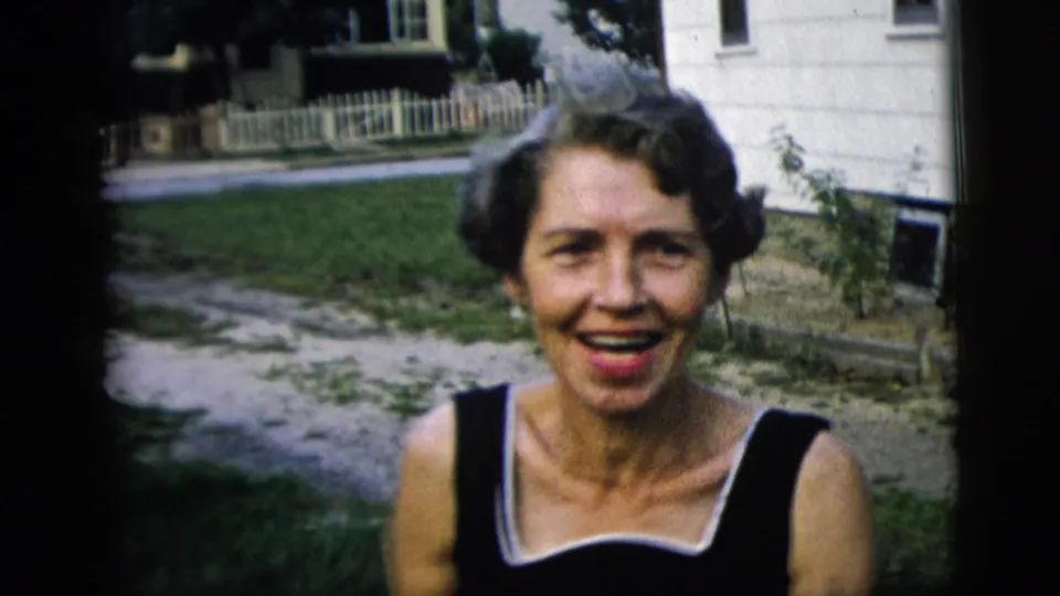 The woman smiles broadly near the camera, with a lawn, pale fence, and residential exterior visible behind her.