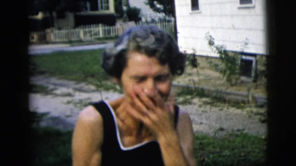 The woman appears bashful or laughing as she covers her mouth in the residential yard, with house siding and path behind her.