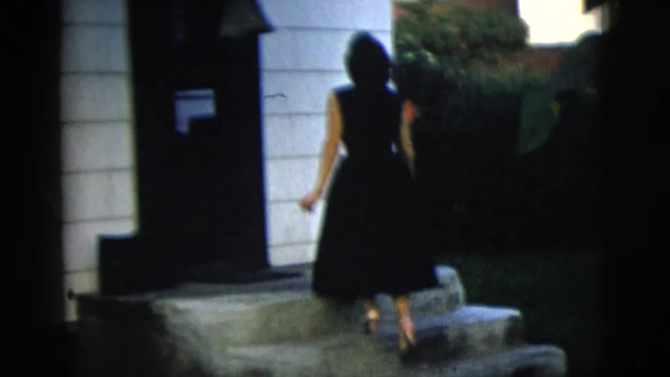 The woman turns away and walks up stone steps toward a doorway, her dark dress visible against the light house siding.