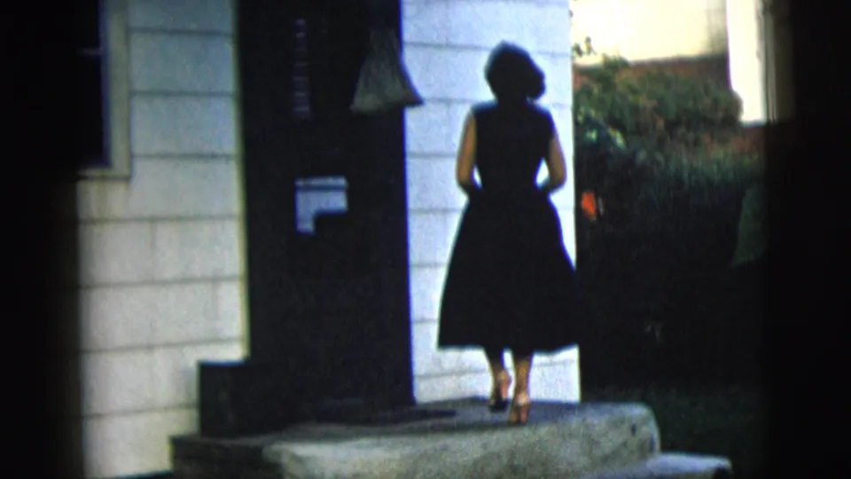 In 1958 New York City home-movie footage, a woman in a black dress stands at a residential entrance with white siding.