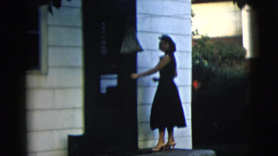 The woman turns beside a dark residential doorway, with white siding and garden greenery visible around the entrance.