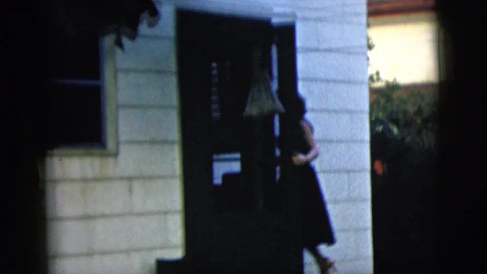 The woman moves into the shadowed doorway, partly obscured by the dark entrance and porch framing.