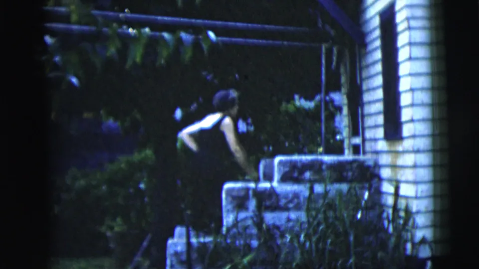 Near the side of the house, the woman leans by stone steps with plants and white siding visible in the frame.
